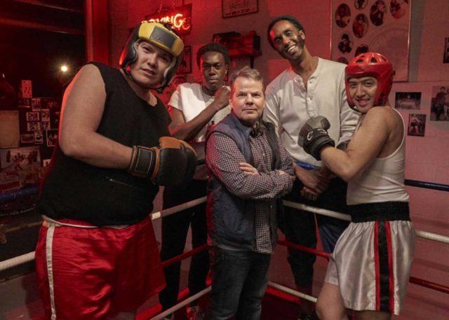 A man stands in a boxing ring in the middle of four men wearing boxing gloves and helmets.