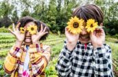 Two smiling children in a garden holding yellow flowers over their eyes.