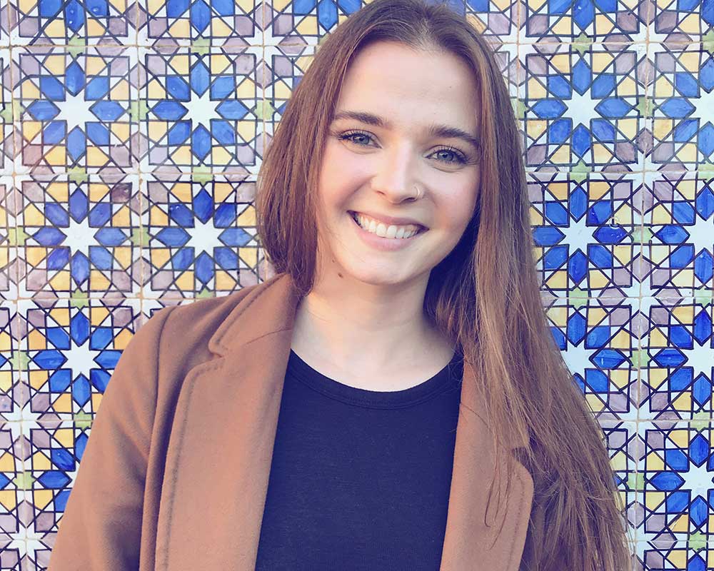 Headshot of women with long dark hair wearing a brown jacket standing in front of a colourful mosaic tiled wall.