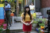 A girl with bangs and long dark hair wearing a yellow t-shirt standing in a backyard.