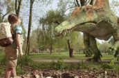 A brown haired girl wearing shorts and a backpack stands looking up to a dinosaur.