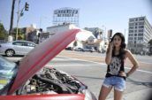 Woman on cell phone standing with hand on her hip in front of a broken down red car.